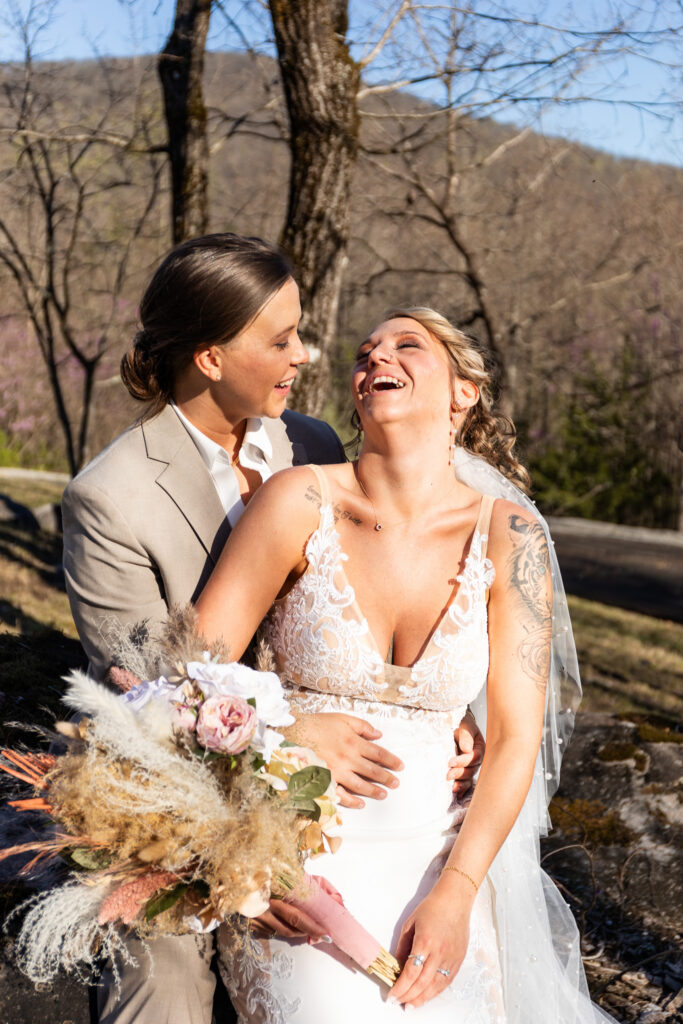 A bride in a lace wedding dress with a pearl-detailed veil throws her head back laughing while being embraced from behind by her spouse in a tan suit jacket. The bride holds a textured bouquet filled with pampas grass, roses, and dried florals as golden afternoon light illuminates the couple during romantic wedding portraits outdoors among rocky terrain and early spring trees.