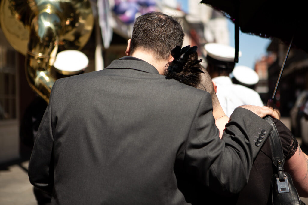 A man in a dark suit wraps his arm around a grieving woman’s shoulders as they walk through the streets of New Orleans during a second line funeral. Brass instruments gleam in the sunlight ahead of them, a reminder that music and community play a vital role in overcoming loss and carrying love forward.