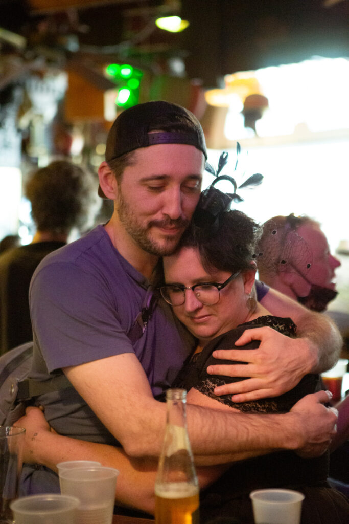 Inside a dimly lit bar, a woman in mourning is embraced tightly by a friend, their arms wrapped around each other in silent understanding. Their connection is a testament to the strength found in community and the deeply personal journey of overcoming loss.