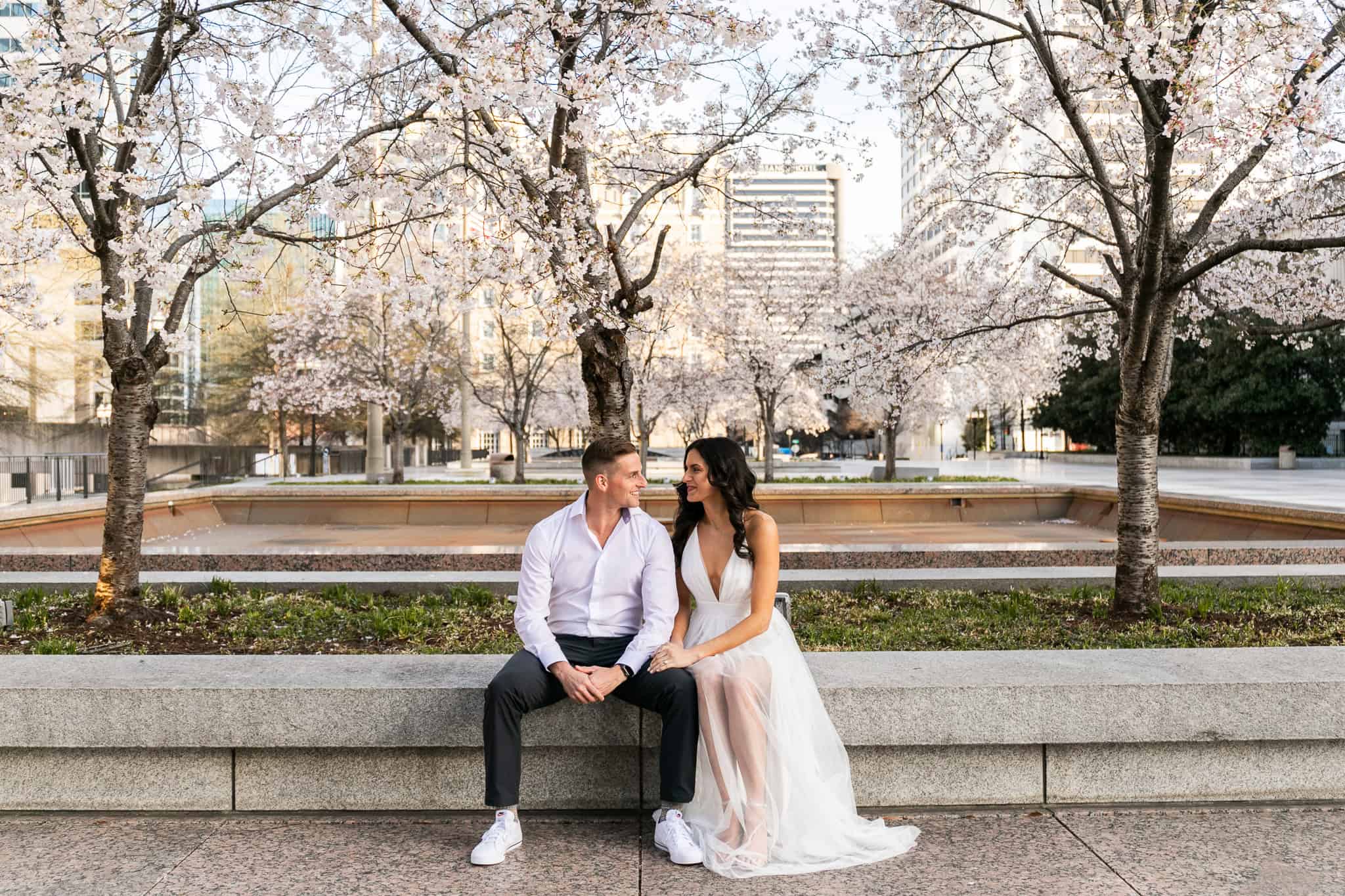 A romantic engagement session in an urban park with cherry blossoms in full bloom. The couple sits closely on a stone bench, gazing into each other's eyes. The woman wears a flowing white dress, and the man is dressed in a white shirt and dark slacks. The pastel pink flowers contrast beautifully against the city skyline