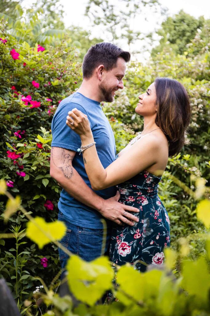 A lush, vibrant outdoor engagement session surrounded by blooming pink flowers. The couple stands closely, gazing into each other’s eyes with soft smiles. The groom wears a casual blue shirt, showcasing a tattoo on his arm, while the bride wears a floral dress, her engagement ring subtly visible as she touches his shoulder