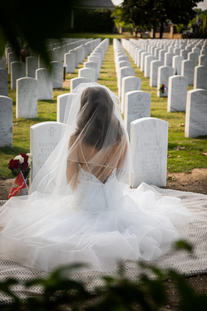 Captured from behind, a bride in an ethereal wedding gown kneels before her fiancé's grave, her veil draping down her back as she faces the endless rows of headstones. The golden light filters through, symbolizing resilience and remembrance. This poignant moment reflects the heartache of loss and the quiet strength of overcoming grief—navigating love, memory, and the future with unwavering grace