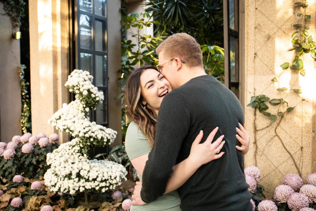 Bathed in soft golden light, a couple embraces inside the Conservatory at Longwood Gardens, surrounded by lush greenery and delicate floral arrangements. 