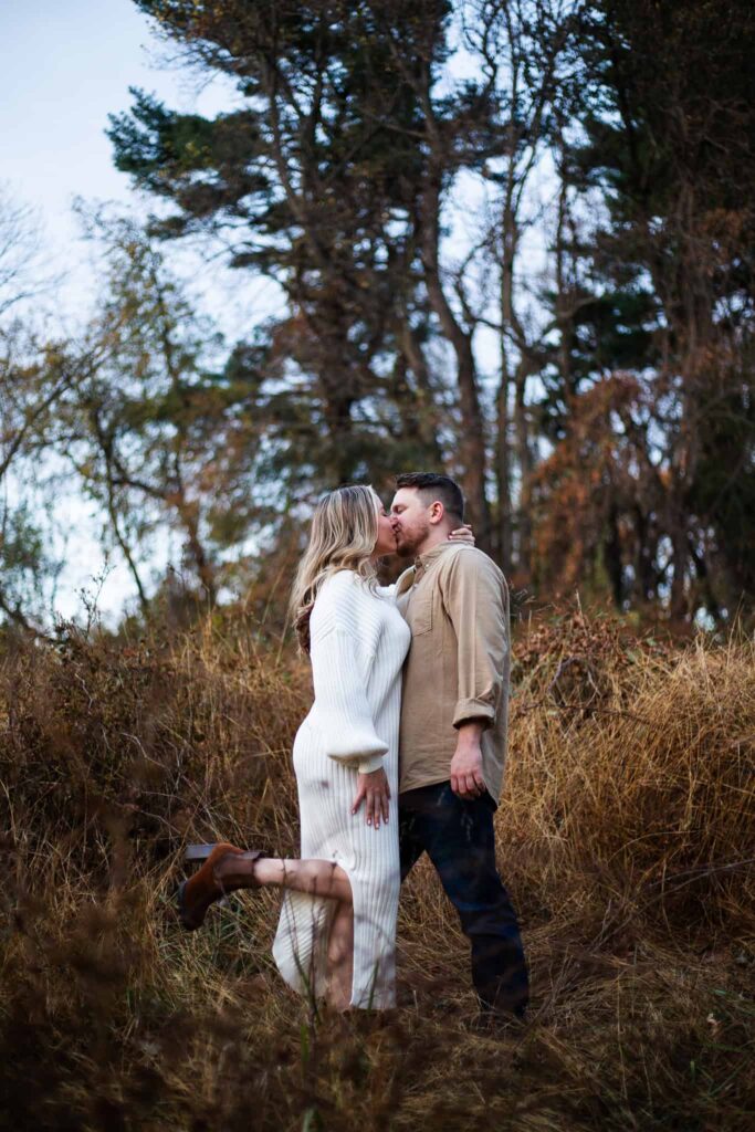 A romantic and moody engagement photo taken in a rustic outdoor setting. The couple stands in a field of tall, golden grass, surrounded by towering trees. The bride, wearing a cozy white knit dress, lifts her leg playfully as she kisses her groom, who is dressed in a casual beige shirt and dark jeans, their love radiating warmth in the cool evening light