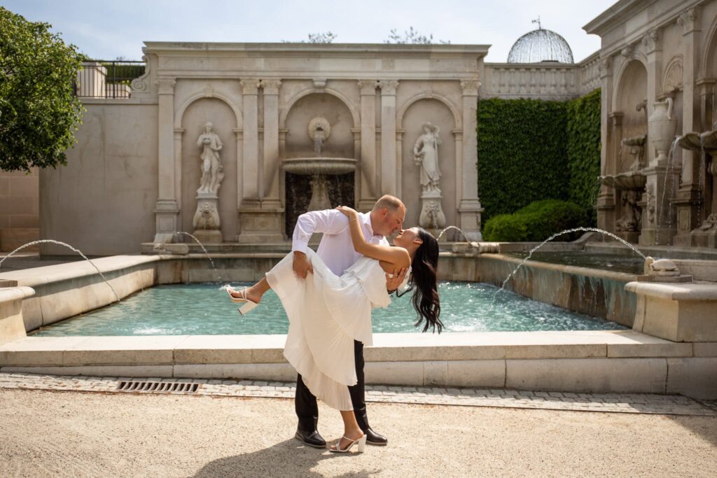 A romantic engagement photo at Longwood Gardens featuring a groom dipping his bride in front of an elegant stone fountain framed by classical sculptures. The soft ripples of the water and grand architecture highlight the timeless beauty of the gardens