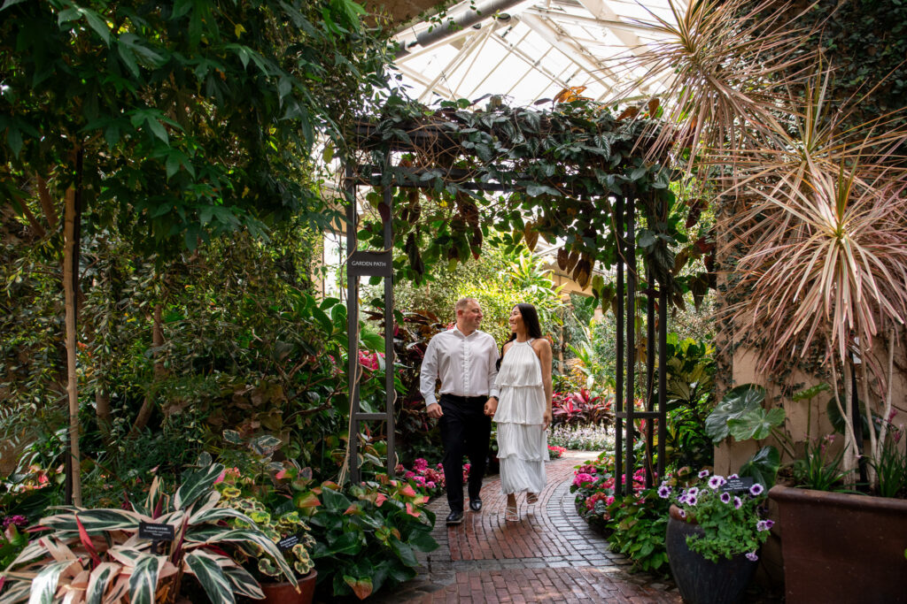 A couple strolls hand-in-hand beneath an ivy-covered archway in the lush indoor gardens of Longwood Gardens. Vibrant flowers in shades of pink and purple line the cobblestone path as they share a quiet moment during their engagement session
