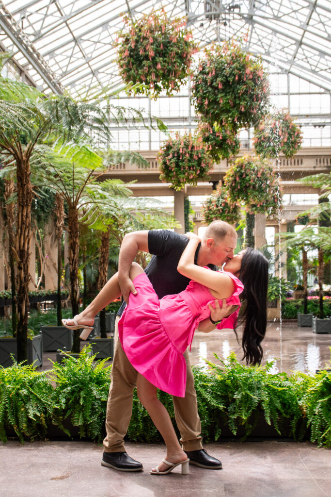 A groom dips his bride in an elegant pose beneath cascading floral chandeliers at Longwood Gardens' conservatory. She wears a bright pink dress that contrasts beautifully with the lush greenery, while a delicate fountain trickles in the background.