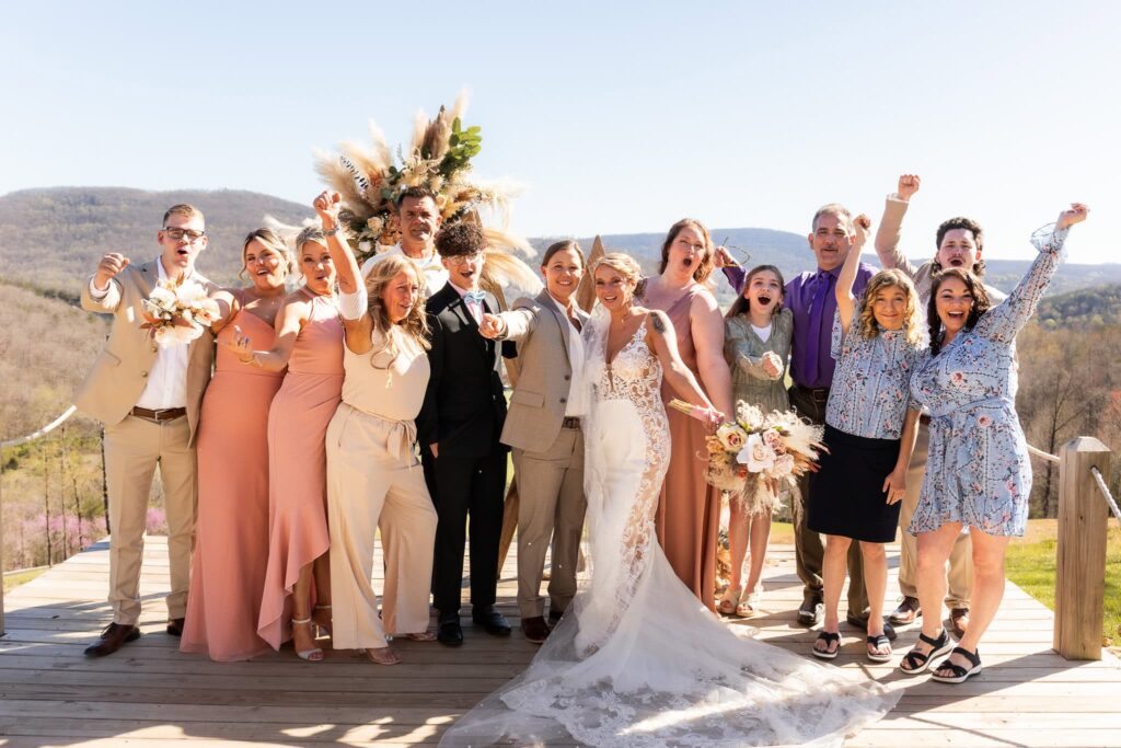 Two Brides cheer on their wedding day with family and friends, dressed in a mix of pastels and neutrals, cheers in celebration against a breathtaking mountain backdrop. 