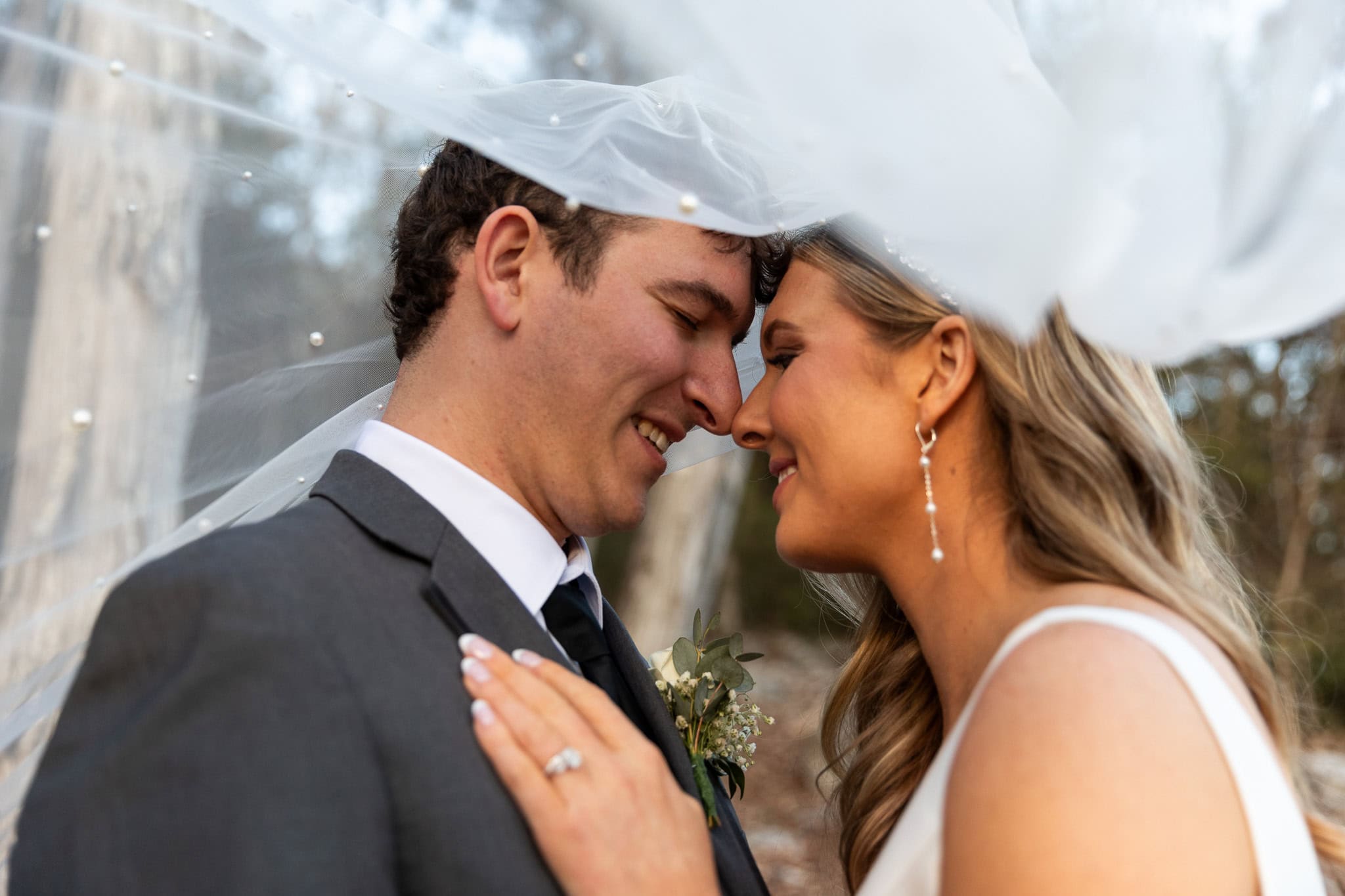 A bride and groom share a tender moment under the bride’s delicate pearl-studded veil, which softly drapes over them like a dreamy canopy. Their noses touch as they exchange smiles, their eyes filled with love. The bride’s hand rests on the groom’s chest, her engagement ring sparkling in the soft light. The groom, wearing a charcoal suit with a crisp white shirt and black tie, looks at his bride with warmth and joy. In the background, blurred trees add a natural, romantic feel, perfectly complementing this intimate wedding portrait