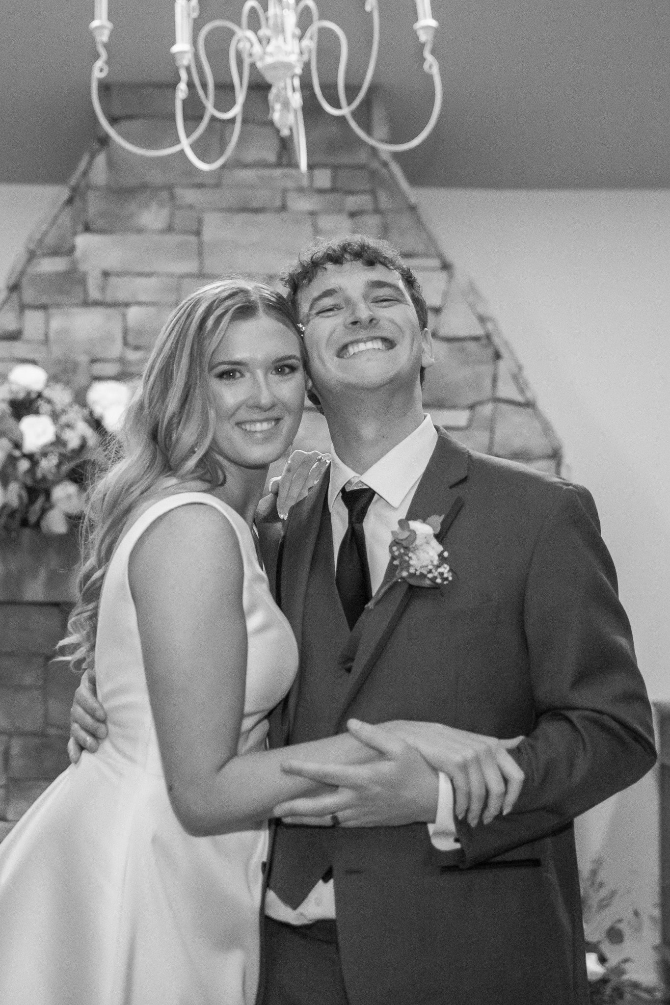 In front of the stone fireplace, the couple embraces, their faces lit with pure happiness. The groom’s broad grin fills the frame as the bride wraps her arms around him, leaning in close. The soft glow from the reception lights highlights their features in this timeless, black-and-white portrait.