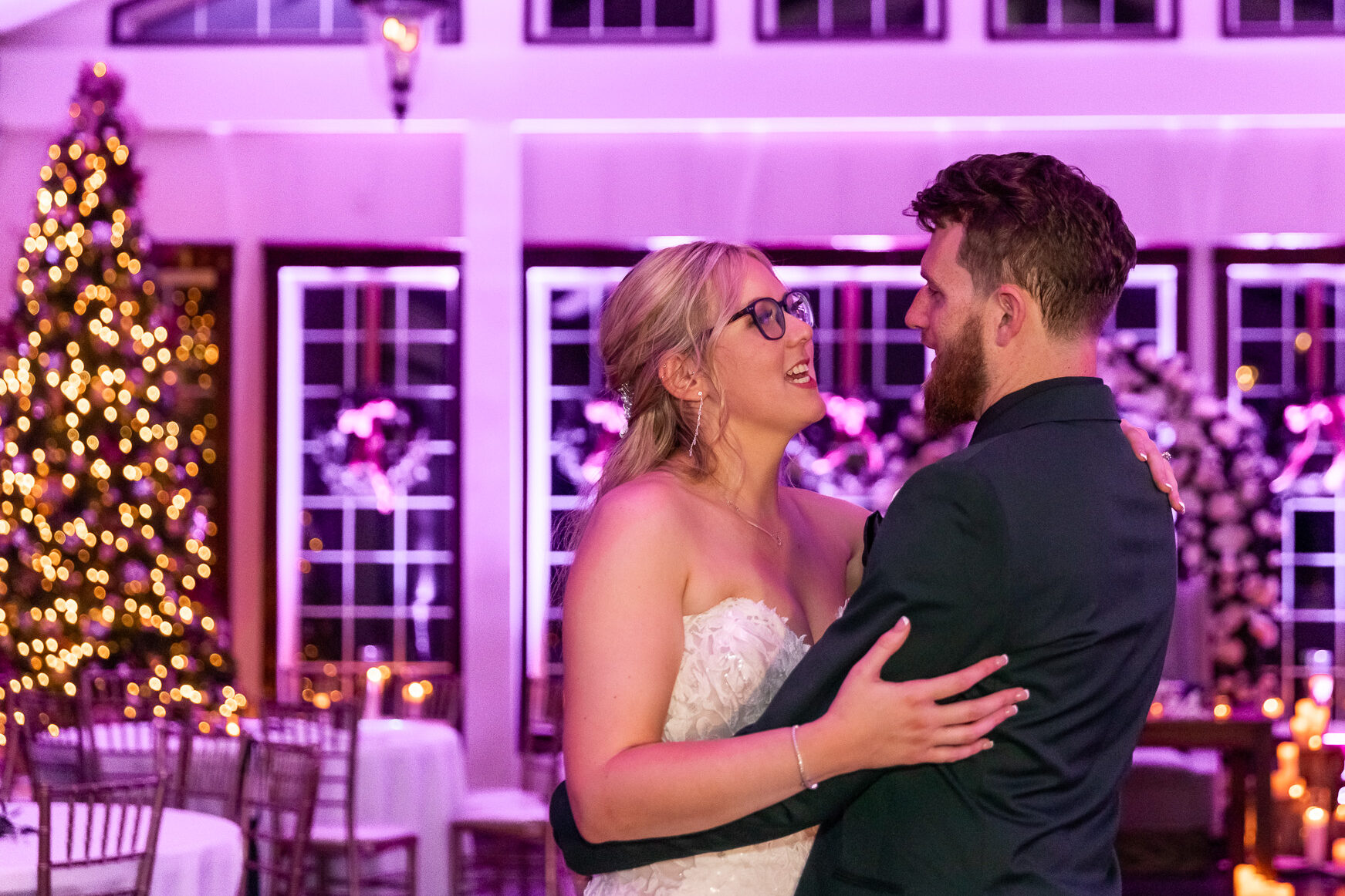 The bride and groom hold each other close on the dance floor, lost in the moment as they sway under the soft glow of twinkling Christmas lights. The Running Deer Golf Club is bathed in a romantic purple hue, with warm candlelight flickering around them. A towering Christmas tree glows in the background, adding to the dreamy atmosphere. Their eyes meet, and their smiles reflect pure joy as they soak in their first dance as a married couple.
