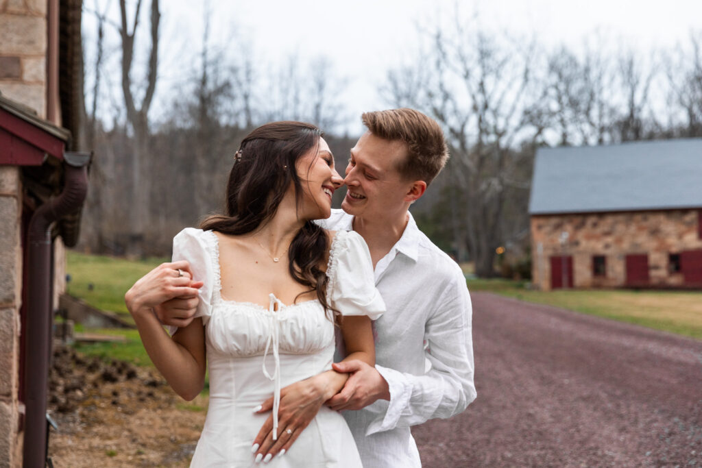 A couple in white outfits share a joyful, nose-to-nose moment on a gravel driveway with a historic stone barn in the background. The woman holds her partner's hands around her waist as they laugh under an overcast sky, the mood playful and romantic.