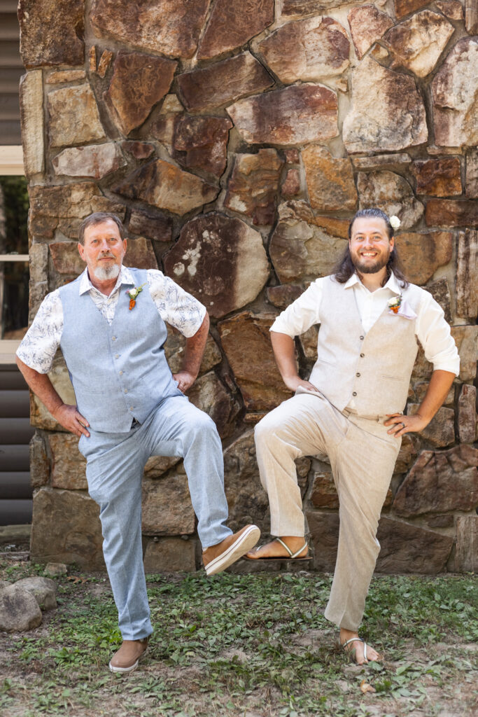a groom dressed in a tan suit with no jacket stands next to his father in a similar attire in baby blue. Both are lifting one foot off the ground "Captain Morgan Style" with their hands on their hips.