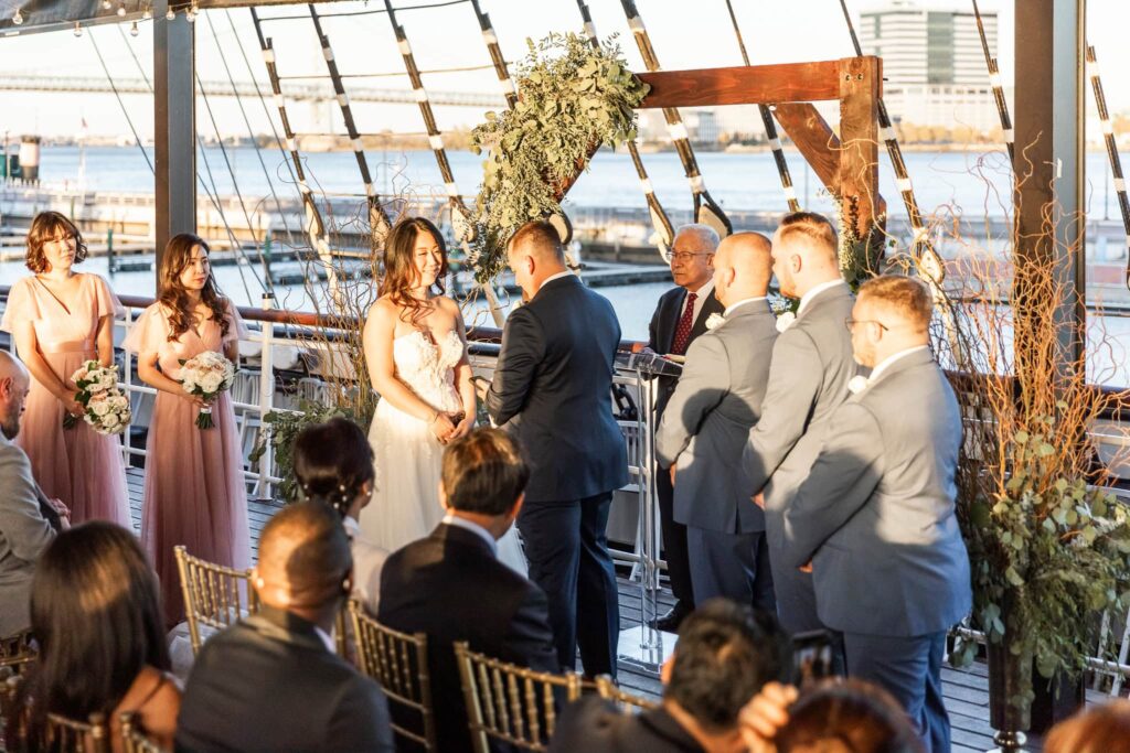 The bride and groom stand before a wooden wedding arch covered in lush greenery as they exchange vows on the Moshulu’s deck. Sunlight filters through the ship’s rigging, illuminating the moment while guests watch intently.