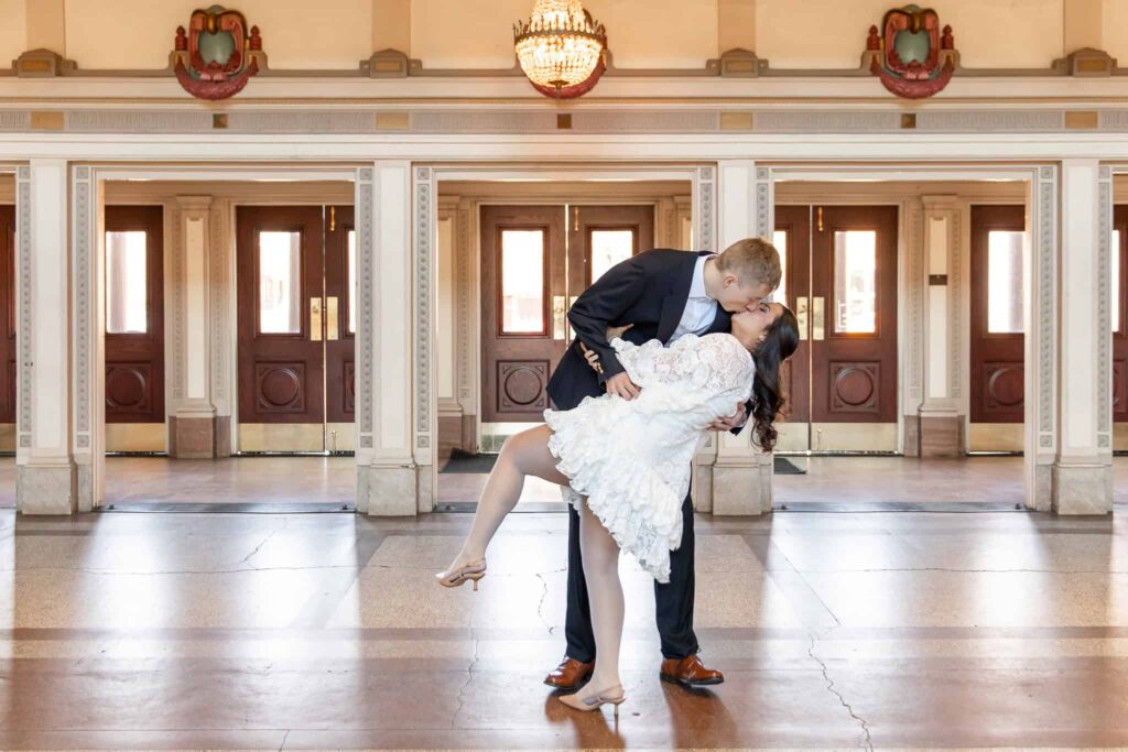 A breathtaking engagement session set in an elegant historic venue, where a couple shares a romantic dip-kiss beneath a grand chandelier. The groom, dressed in a navy suit and brown leather shoes, leans in to kiss his fiancée, who wears a short, ruffled white lace dress and nude heels. Her leg lifts playfully as she embraces him, showcasing their effortless chemistry. The warm wooden doors and intricate architectural details frame this timeless moment