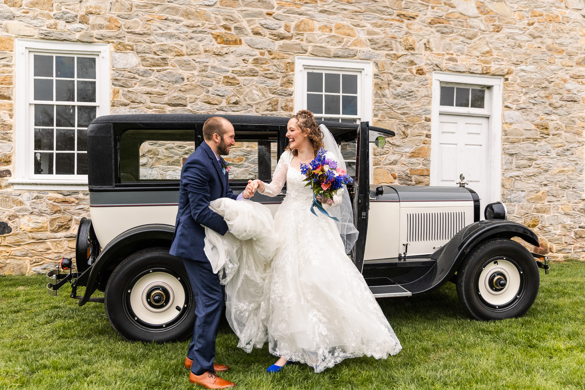 The groom helps the bride out of the vintage car, holding the hem of her gown with one hand and her arm with the other. They laugh together, sharing a joyful exchange outside Haldeman Mansion’s stone facade.