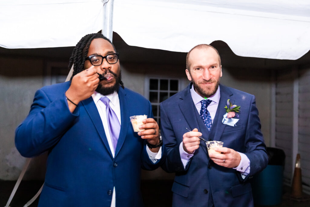 The groom and a groomsman dig into cups of dessert under the reception tent, each with a spoonful mid-air, enjoying a relaxed late-night treat. A candid, lighthearted memory from their Haldeman Mansion Wedding.