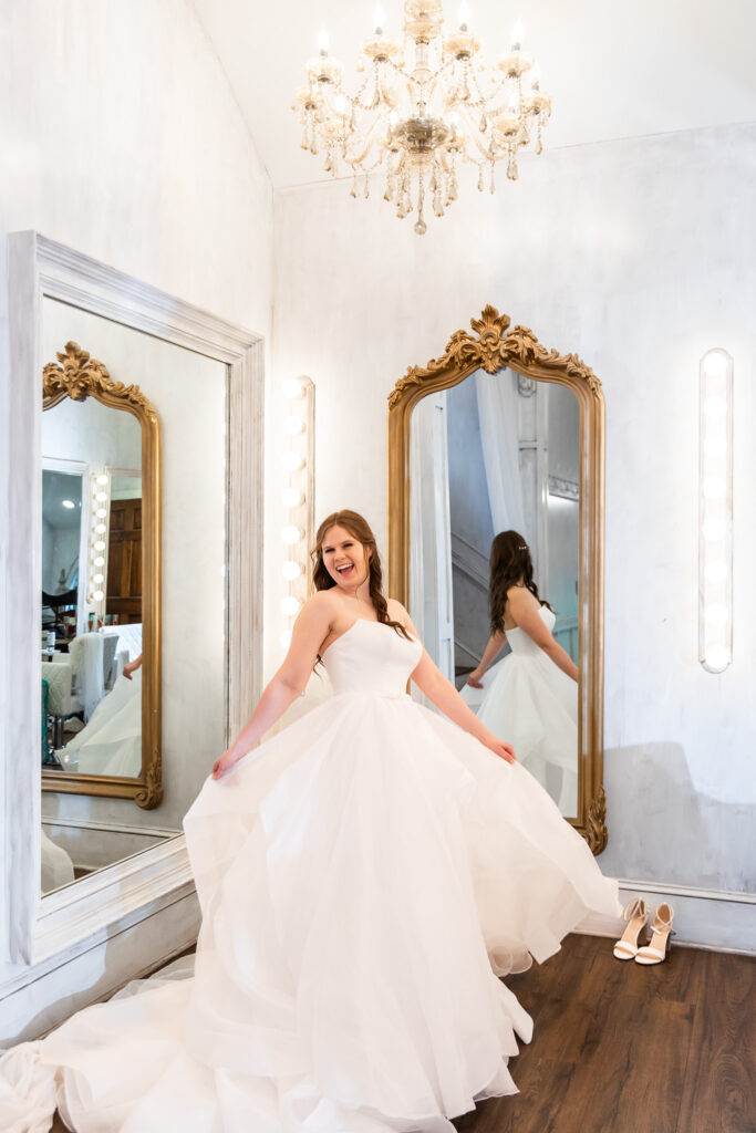 A joyful moment of the bride twirling in her layered ballgown in front of ornate gold mirrors and chandeliers inside the getting-ready room at Highlands Chapel.