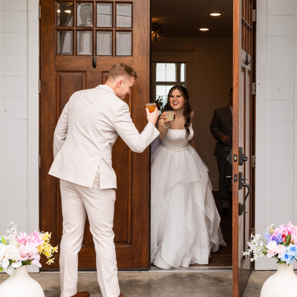 The groom raises a cocktail glass as he approaches the chapel doorway, where the bride, glowing in her gown, greets him with a joyful toast just outside the entrance to the Chattanooga wedding chapel.