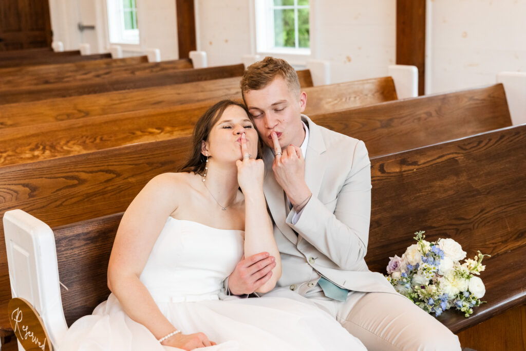 A playful moment between the newlyweds as they sit side by side in a wooden pew, holding up their wedding bands with puckered lips, sharing a quiet laugh in this romantic and spontaneous capture inside a Chattanooga wedding chapel.