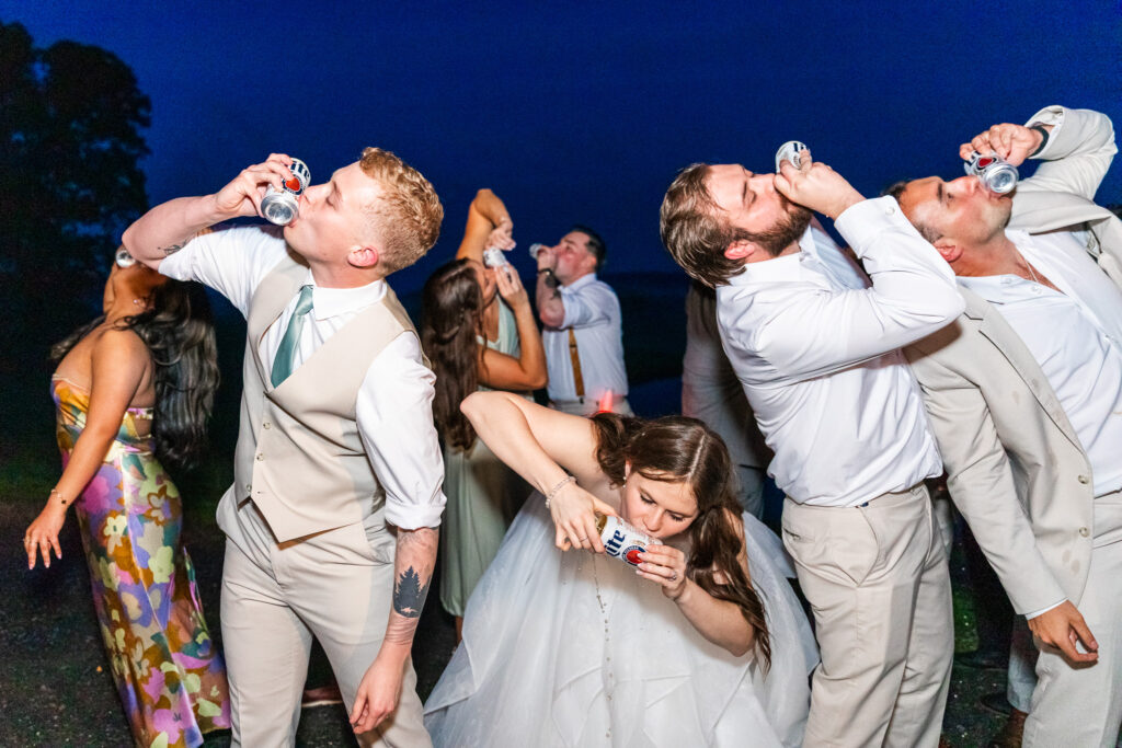 Late in the evening, the couple and friends shotgun beers under a navy-blue sky. The bride is bent forward in her voluminous gown, mid-drink, while the groom and others tilt their heads back with cans in hand. The moment is loud and celebratory, full of energy.