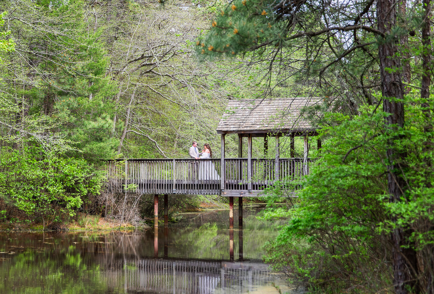 From a distance, the couple appears small and serene, framed perfectly on a wooden bridge that stretches over glassy water. The reflection of the trees and structure below them made this Medford, NJ wedding scene feel almost storybook-like.