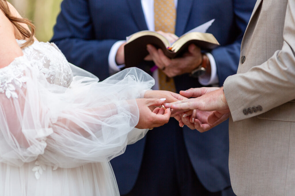 A close-up of the couple’s hands during the ring exchange. The bride, in a sheer off-the-shoulder sleeve, gently slides a wedding band onto the groom’s finger while the officiant holds an open Bible in the background. This emotional moment was one of many that made this Lakeside at Medford wedding feel deeply rooted in love and intention.