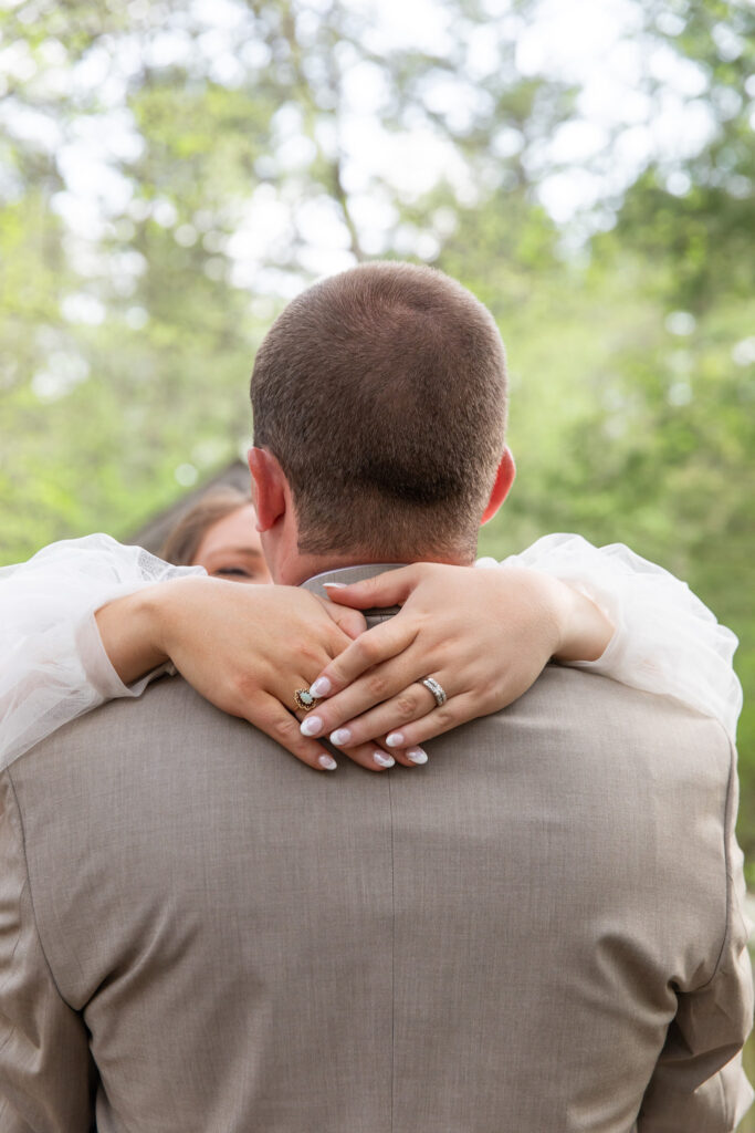 A quiet, intimate moment as the bride’s arms wrap around the groom’s shoulders from behind, her hands resting gently on his back. Her engagement ring and wedding band sparkle against the soft tulle of her dress. This sweet detail captures the intimacy and serenity that define a Lakeside at Medford wedding celebration.