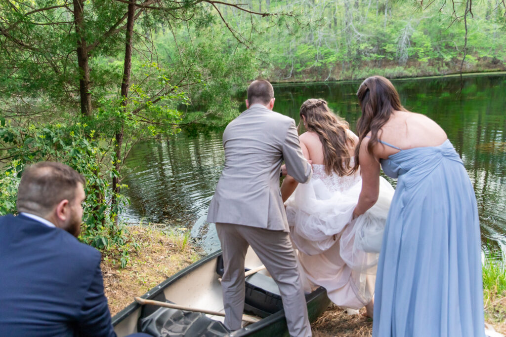 The bride is assisted into a canoe by the groom and bridesmaid while another groomsman steadies the boat. They are at the edge of a wooded lake, with the calm water reflecting the trees around them. The moment is both cautious and exciting, filled with laughter and helping hands.