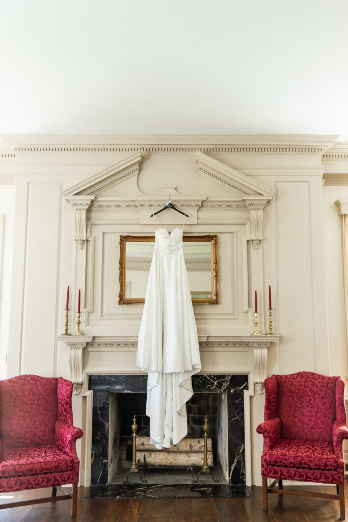 A strapless white wedding gown with a sweetheart neckline hangs from a black hanger centered above an ornate fireplace at Welkinweir Estate. The creamy paneled wall and antique mirror behind it add classic charm, while two red velvet armchairs and brass candlesticks frame the scene, capturing the elegant, historical ambiance of this Welkinweir Estate wedding.