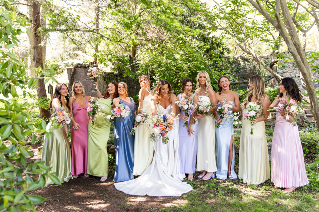 The bride and her ten bridesmaids beam with joy in a shaded grove, wearing mismatched pastel gowns that echo the wedding’s wildflower palette. This lively bridal party moment is full of color, laughter, and the natural beauty of a central PA wedding.