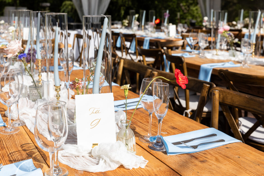 A long wooden reception table at Welkinweir Estate is set with pale blue napkins, gold-accented glassware, hand-lettered table numbers, and tall candlesticks in glass hurricanes. Wildflower buds in small vases complete the romantic garden theme of this Welkinweir Estate wedding.