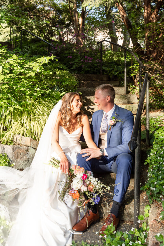 Seated on a stone staircase nestled in greenery, Olivia and Noah laugh together with a vibrant bouquet in her lap at their Welkinweir Estate wedding. The golden hour light brings warmth to their relaxed moment.