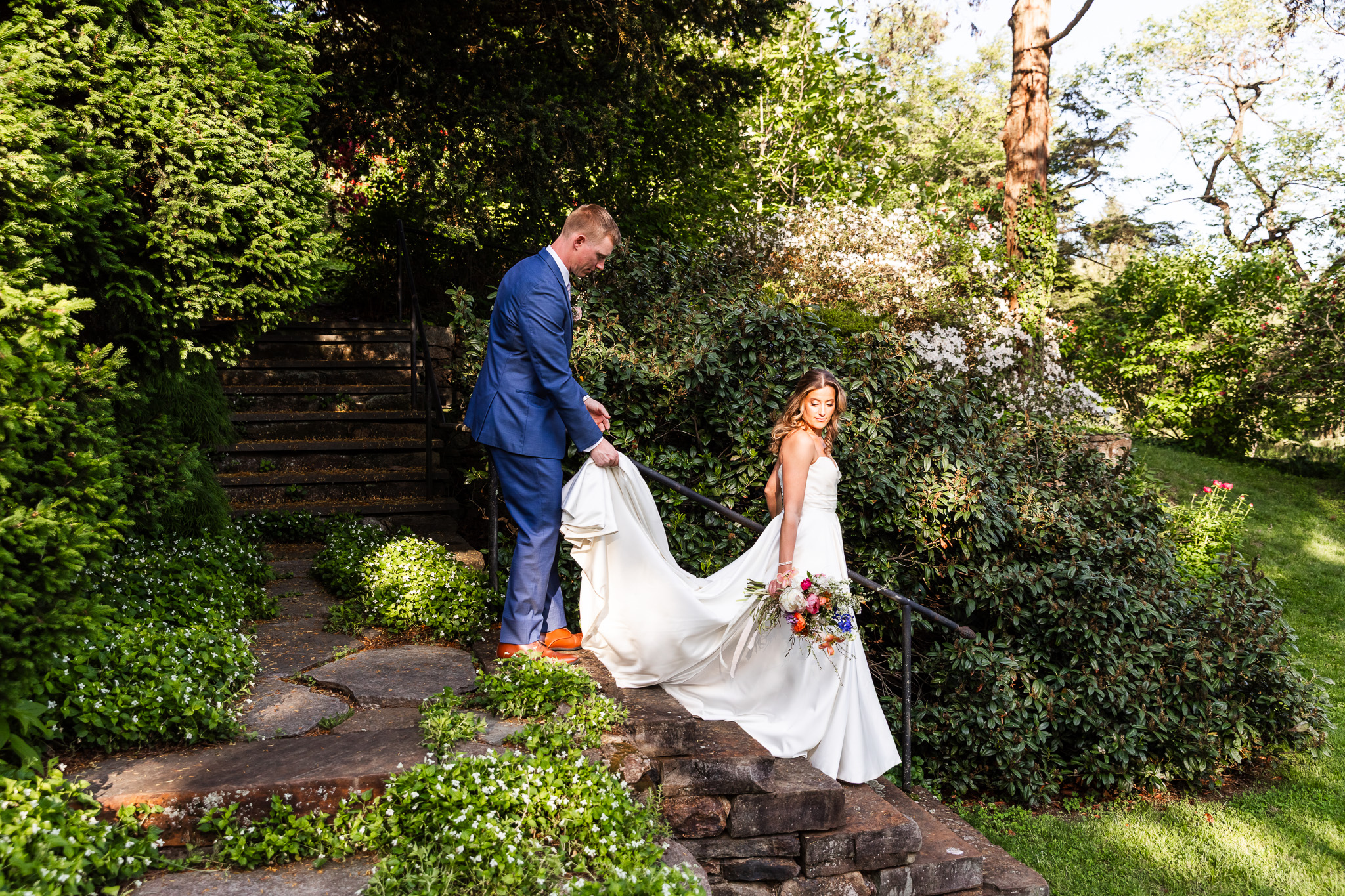 The groom gently holds the bride’s train as she walks down stone garden steps surrounded by lush greenery and blooming white flowers. The bride glances back gracefully while holding a bouquet of wildflowers, framed by a soft glow of afternoon sunlight