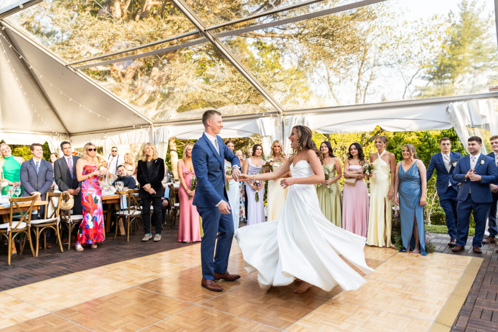 The bride twirls across the dance floor with her gown sweeping out around her as the groom leads their first dance. Guests in colorful dresses and suits smile in the background beneath a clear tent filled with warm golden sunlight at a Pottstown wedding venue.
