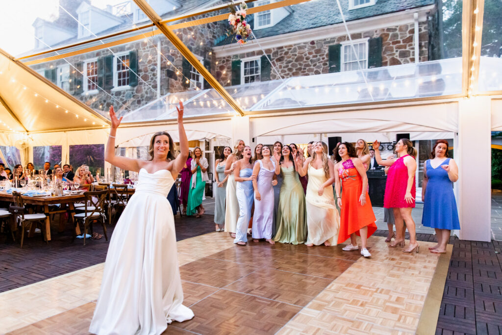The bride tosses her colorful bouquet into the air beneath a clear tent as a group of excited women in pastel and bold dresses gather behind her, a joyful moment that captures the energy of central PA weddings.

