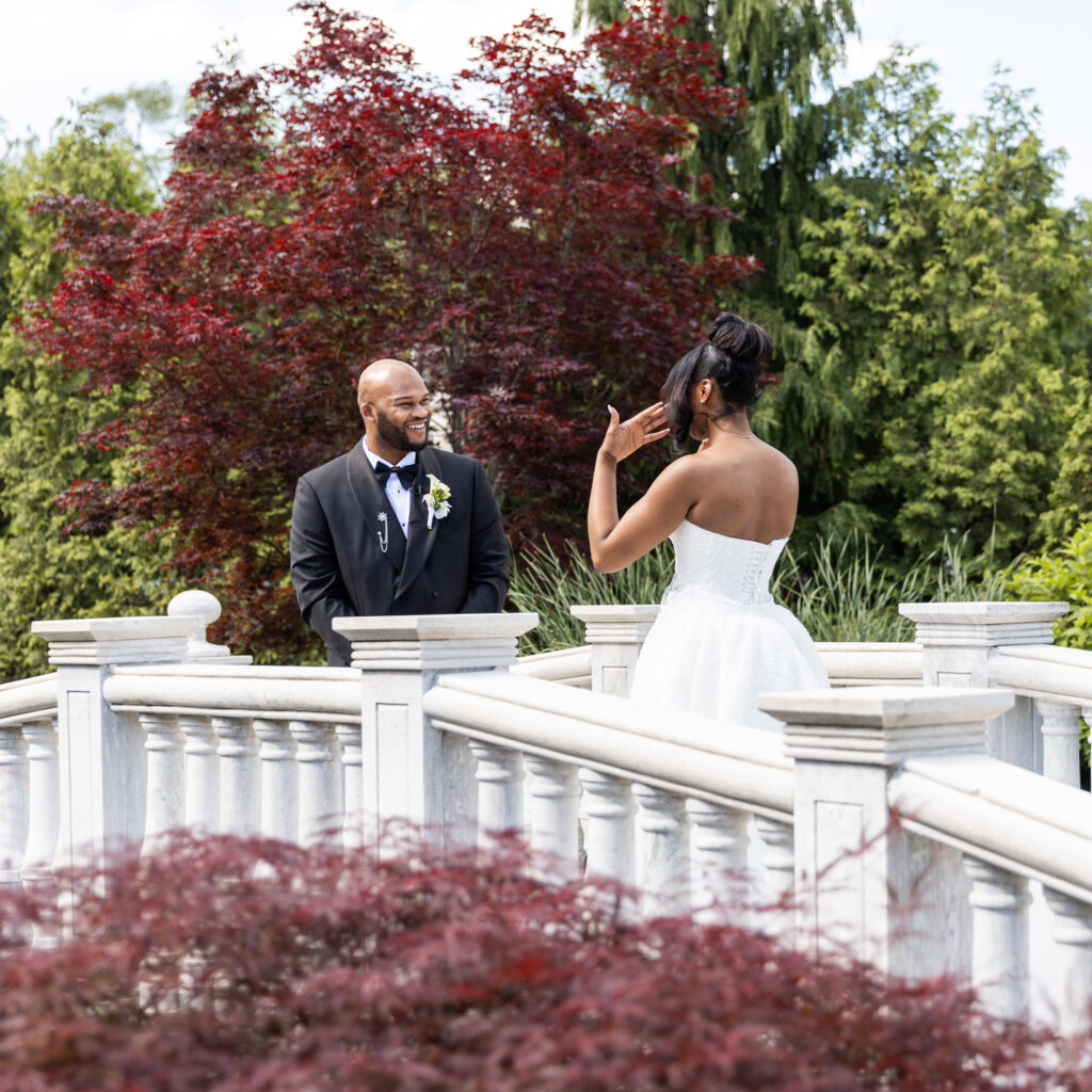 On an outdoor marble bridge surrounded by manicured trees and deep red foliage, Isaiah smiles wide as Zer, in her wedding dress, wipes a tear from her face. The emotion of their first look during their Merion wedding is palpable.