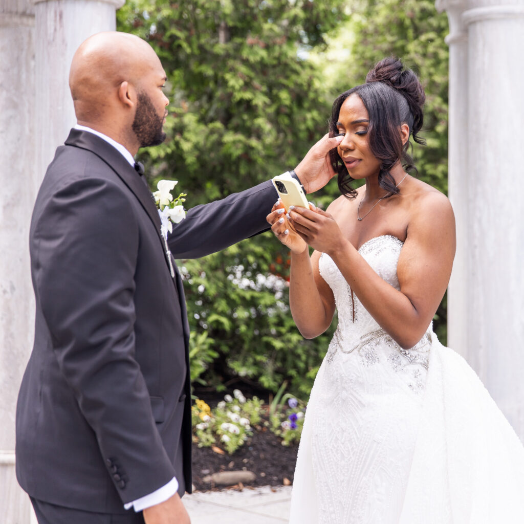 The Merion Wedding moment during private vow exchange as the couple stands near the grand white gazebo, emotional and connected beneath the open sky, the groom wipes a tear from the bride's eye as she reads her vows from her phone.