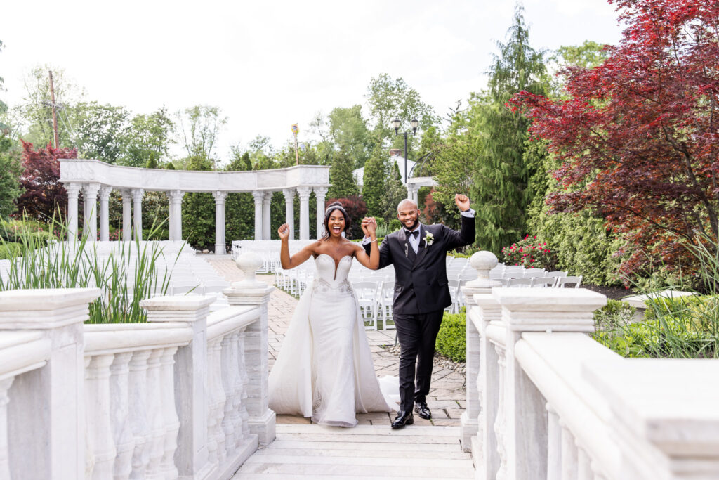 The bride and groom walk hand-in-hand across a white marble bridge outside The Merion, smiling at each other with joyful anticipation. The bride’s strapless gown features intricate beading and a dramatic train, while the groom wears a black tuxedo with a white boutonniere. Behind them, stone columns and lush greenery frame the pathway, with vibrant red foliage adding a pop of color to the garden scene at The Merion.