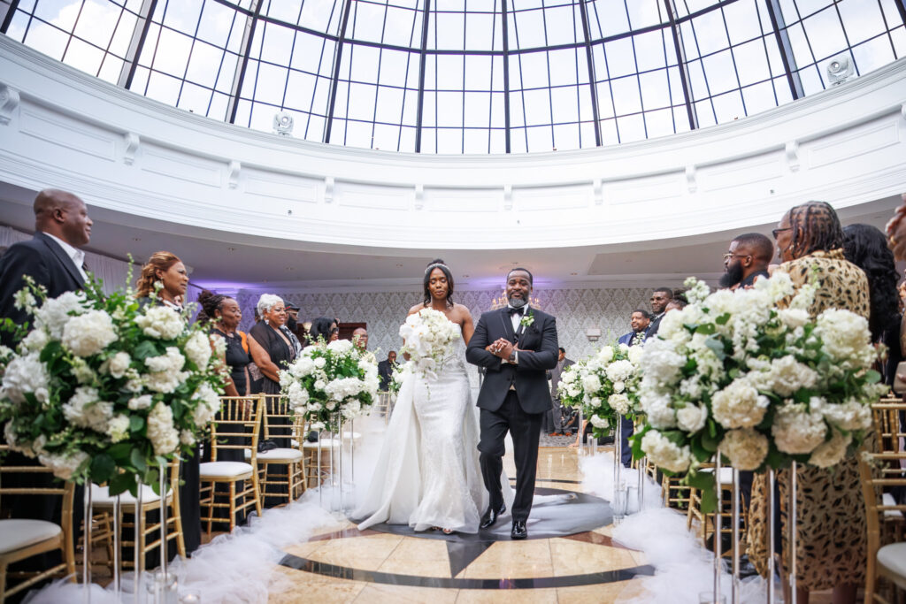 Zer, holding a cascading bouquet of white orchids and roses, walks arm-in-arm with her escort down the aisle. The skylight above floods the space with natural light, and guests stand watching as she approaches the altar during this South Jersey wedding.