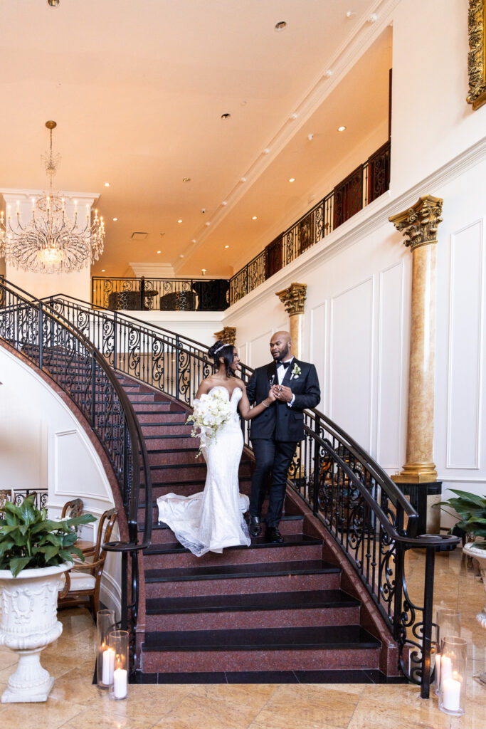 The couple descends the grand curved staircase inside The Merion, holding hands, with the bride carrying a cascading white bouquet under an ornate chandelier, capturing a timeless wedding at The Merion moment.
