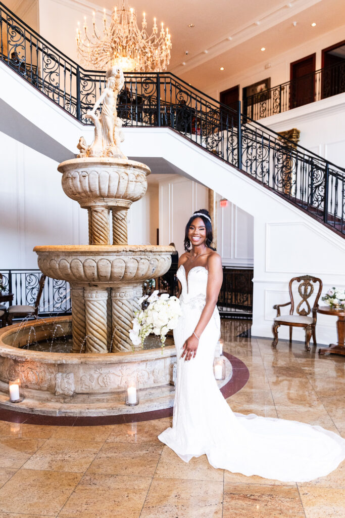 The bride stands inside The Merion beside the same tiered stone fountain, holding her white bouquet and smiling, with a grand chandelier and spiral staircase behind her — a classic indoor bridal portrait from a South Jersey wedding.