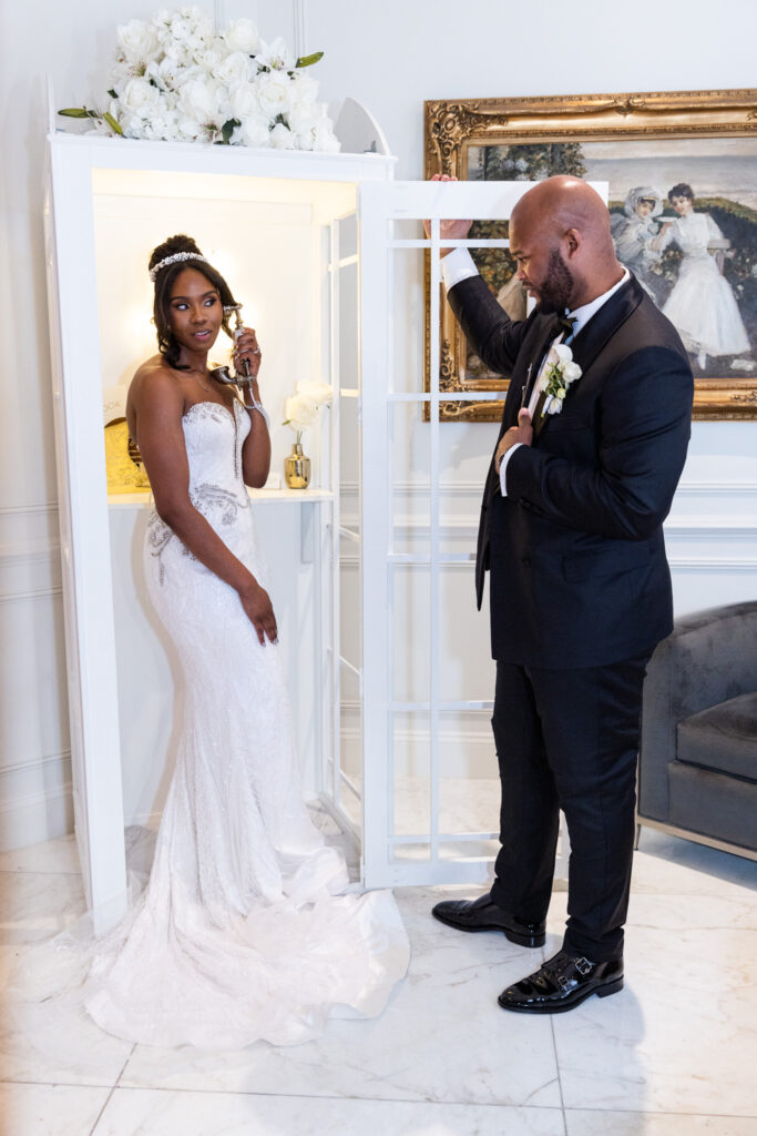 The bride poses with a vintage-style phone inside a white guestbook phone booth adorned with white roses, while the groom playfully leans on the door frame during their wedding at The Merion.