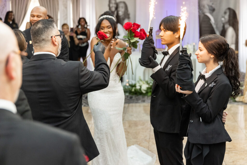 The bride receives a bouquet of red roses with a surprised and delighted expression as she stands near the center of the dance floor. A man in a tuxedo offers her a single rose while two attendants in black gloves and bowties hold bottles with sparklers shooting from the tops. The room is glowing with warm light and celebratory energy, with the bride at the heart of the action, her smile lighting up the scene.