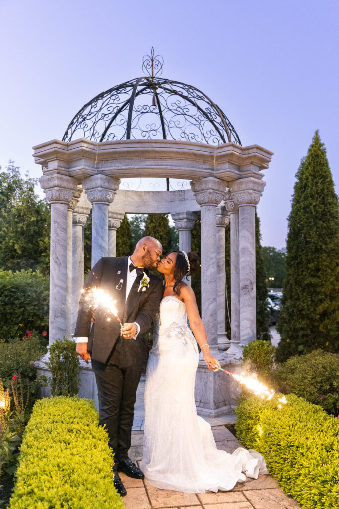 Outside under evening light, the couple kisses in front of the stone-columned gazebo while holding lit sparklers, ending their South Jersey wedding day with joy and romance.