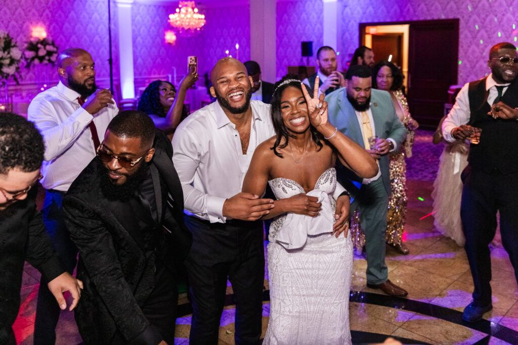 The bride and groom, surrounded by friends, share a laugh and hug on the packed dance floor at The Merion. The bride holds up a peace sign while the groom beams with joy behind her.