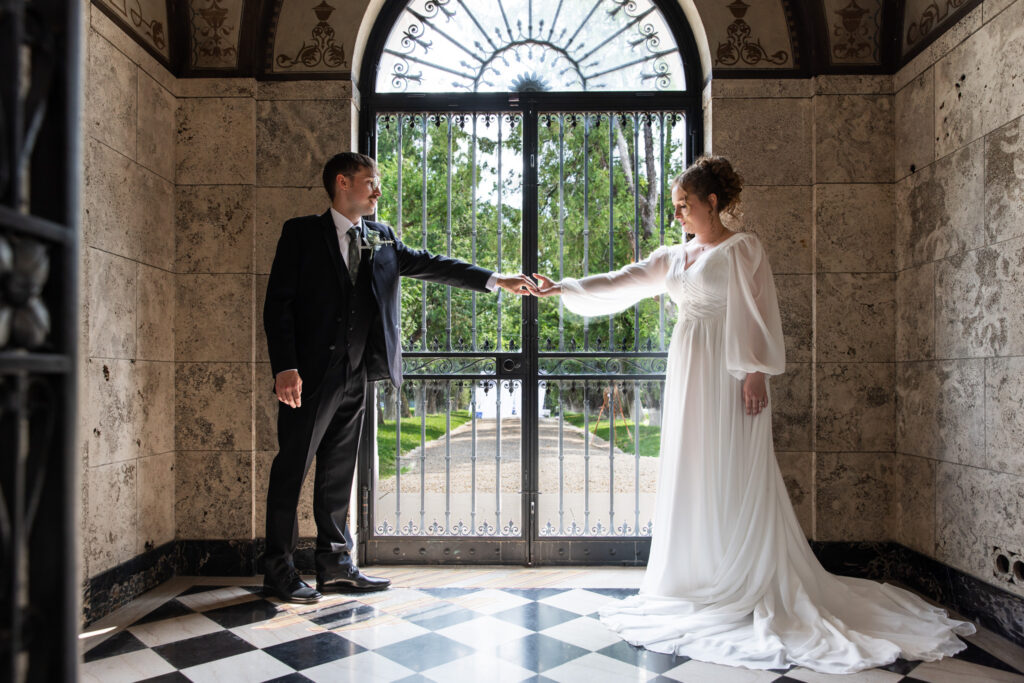 Inside the marble vestibule of the Candoro Marble Building, the couple extends their arms toward each other, fingertips touching. Light pours through the ornate wrought-iron doors behind them, illuminating their formal attire and the checkered black-and-white floor. An artistic portrait full of symmetry and elegance from this Candoro Marble wedding.
