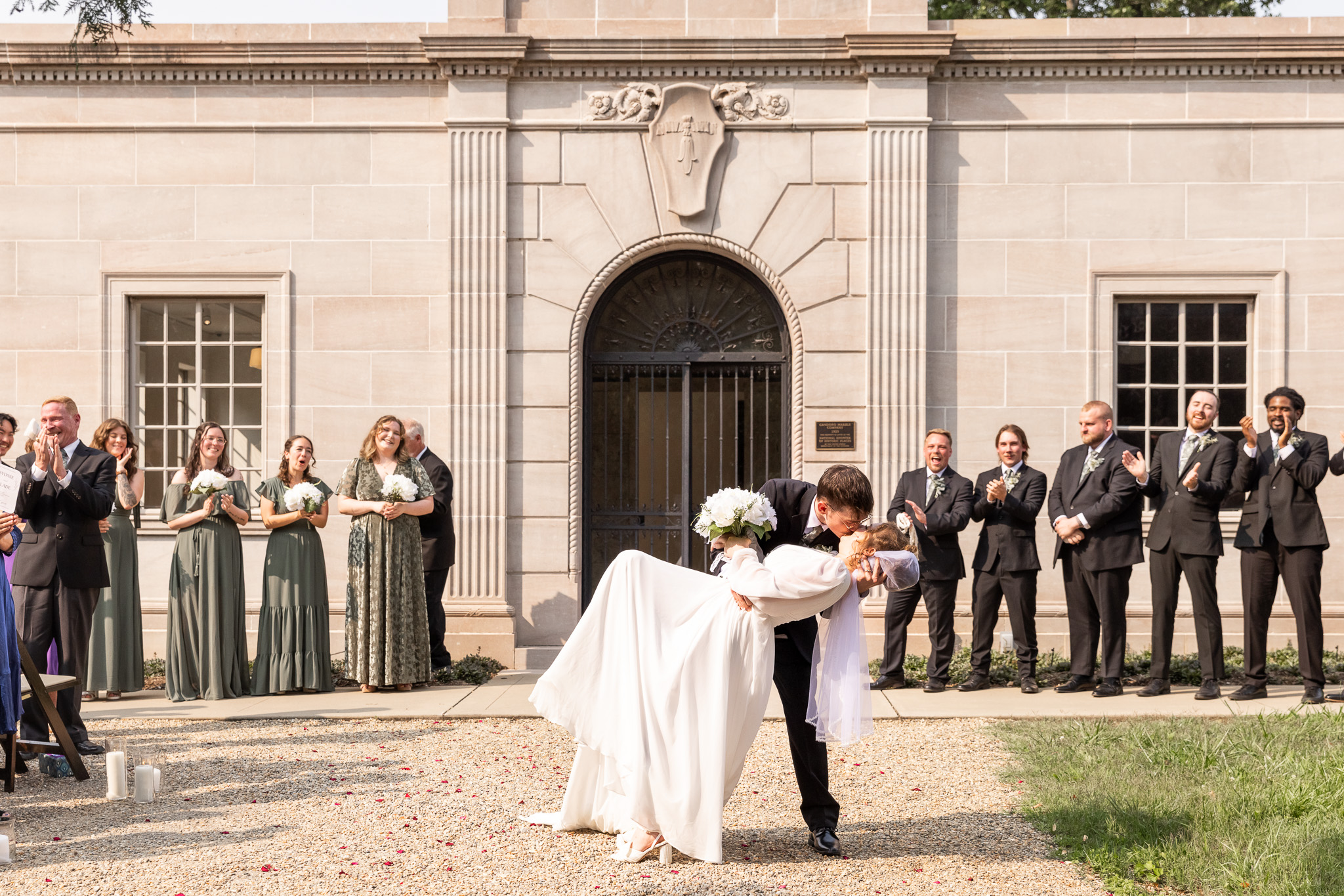 With the Candoro Marble Building as their backdrop, the groom dips his bride in a sweeping kiss as the wedding party cheers in the background. Bridesmaids in sage green dresses and groomsmen in classic black suits line the facade, clapping and smiling. The romantic gesture and classical venue architecture highlight the charm and excitement of this Candoro Marble Wedding celebration.