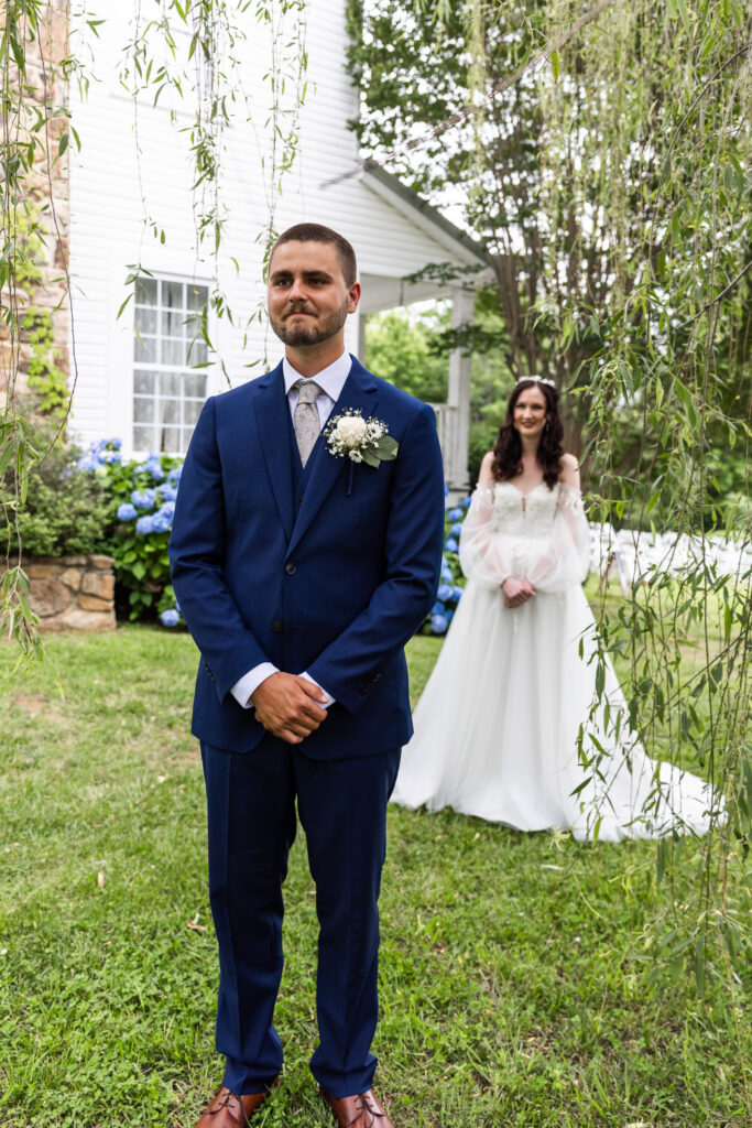 A groom stands with emotion on his face, eyes brimming and lips pressed together, as he waits for the bride to approach for their first look. He wears a dark blue suit with a white boutonniere and stands beneath hanging willow branches, with the bride in the background walking toward him in a flowing gown.