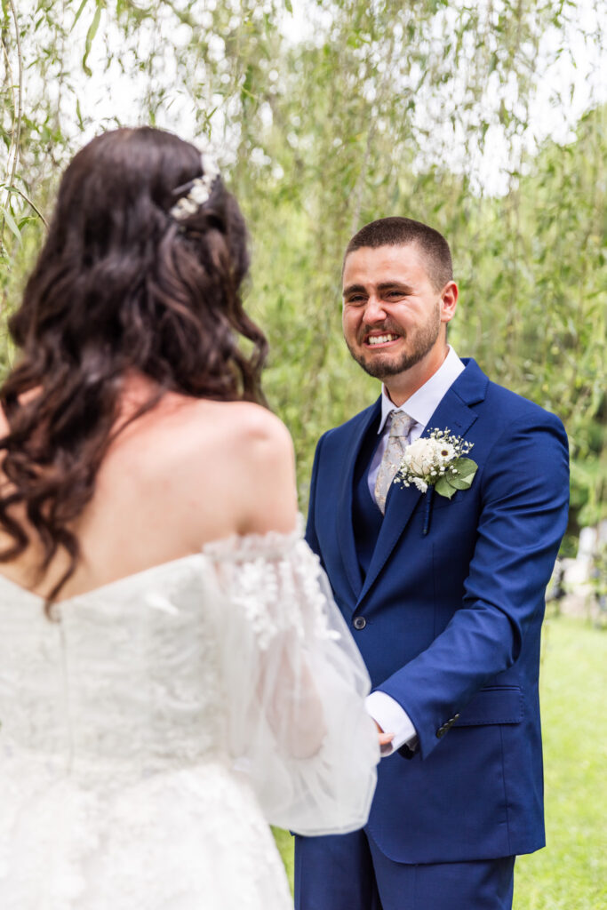 The groom gazes at the bride with misty eyes and a heartfelt smile as they hold hands during their first look. He’s dressed in a navy blue suit, and she wears a white gown with romantic tulle sleeves. They are surrounded by soft green willow trees swaying around them.